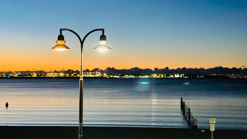 Illuminated street lights by sea against sky at sunset