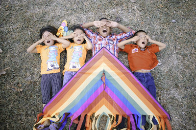 Directly above shot of siblings with hands on eyes and mouth open lying over land with colorful kite