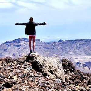 Rear view of woman arms outstretched standing on rock against sky