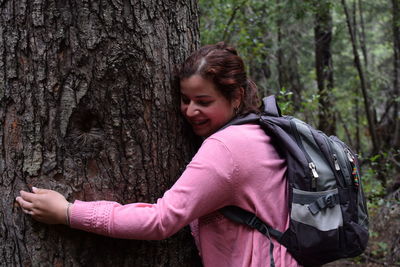 Side view of smiling woman hugging a tree in forest