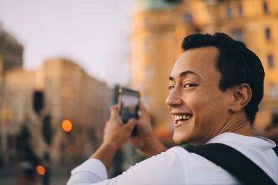 Smiling man looking over shoulder while photographing through phone in city