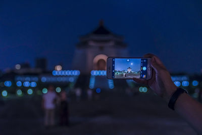 Cropped hand photographing illuminated built structure at night