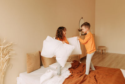 Happy kids brother and sister in pajamas playing fighting with pillows in a cozy bedroom at home