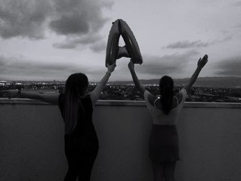 Rear view of friends holding toy on building terrace against cloudy sky