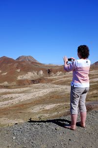 Woman photographing mountains through mobile phone against clear blue sky