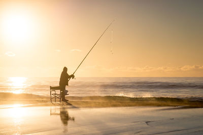 Man fishing in sea against sky during sunset