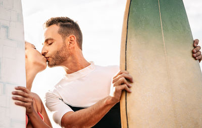 Couple kissing while holding paddleboard against sky