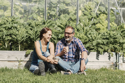 Happy farmers enjoying coffee break while sitting on grass against greenhouse