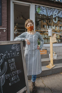 Female store owner with hand on hip standing by blackboard against organic store during covid-19