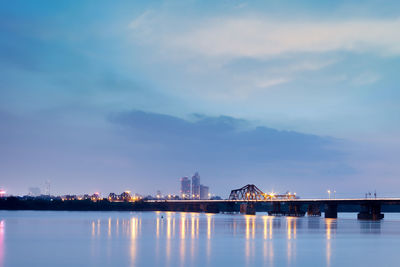 Illuminated city by river against sky at dusk