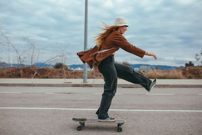 Side view of happy young female millennial in stylish outfit and hat doing trick on skateboard while riding on asphalt road against cloudy sky in countryside