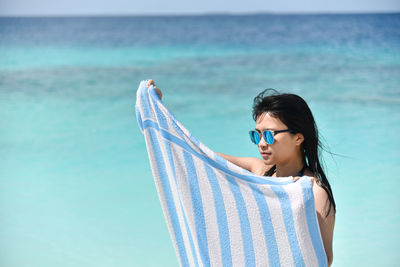 Young woman looking away at sea while holding towel against sky