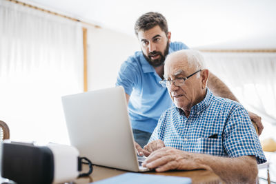 Adult grandson teaching his grandfather to use laptop