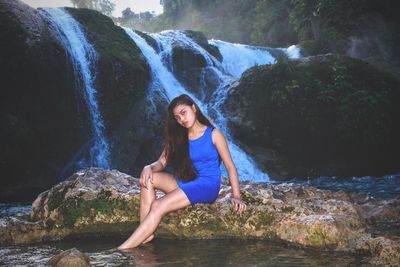 Portrait of beautiful woman sitting on rock formation against waterfall in rainforest