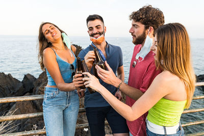 Cheerful friends standing against sea while holding beer