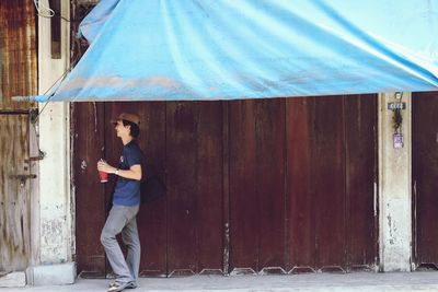 Side view of man with drink looking away while standing against closed wooden door