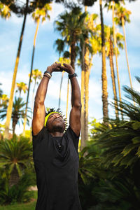 Confident young african american male athlete in trendy sportswear stretching arms while training in park with green tropical trees