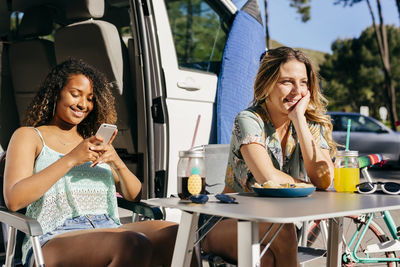 Cheerful women enjoying drink and nachos while sitting out of van in camping using smartphone