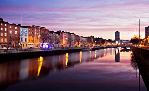 Illuminated buildings by river against sky at sunset