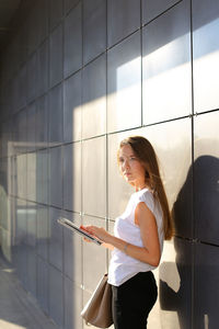 Young woman using digital tablet standing by wall