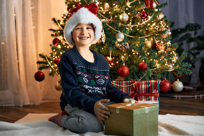 A happy boy in a santa claus hat opens a box with a christmas present