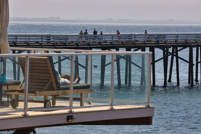 People sitting on railing by sea against sky