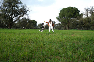 Young lady in casual outfit standing on green grass and trowing ball to black and white obedient spaniel in summer park