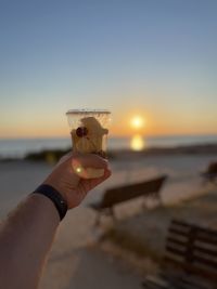 Close-up of hand holding ice cream at beach against sky during sunset