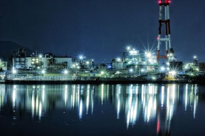 Illuminated factory by river against sky at night