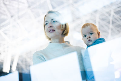 Thoughtful young mother looking trough window holding his infant baby boy child while waiting to