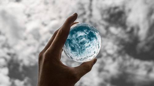 Cropped hand of person holding crystal ball against cloudy sky