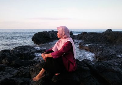 Woman sitting on rock at beach against sky