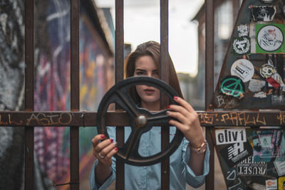 Portrait of young woman holding steering wheel while standing by metallic gate