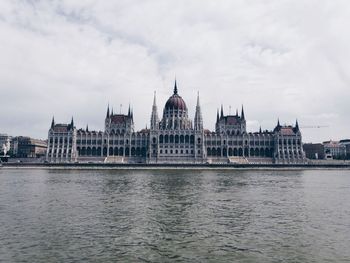 Hungarian parliament building by river against cloudy sky