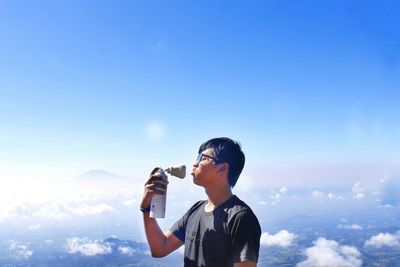 Young man making face while holding drink bottle on mountain against sky