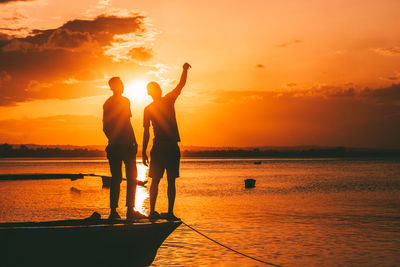 Silhouette of two friends with arms outstretched on beach against sky during sunset