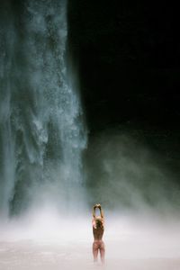 Rear view of woman in bikini standing against waterfall