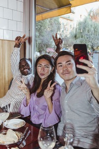 Male and female friends doing peace sign while taking selfie on smart phone at bar