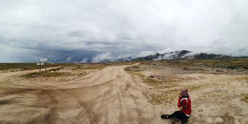 Side view of woman sitting at desert against cloudy sky