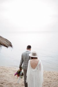 Rear view of romantic newlywed couple walking at beach against sky