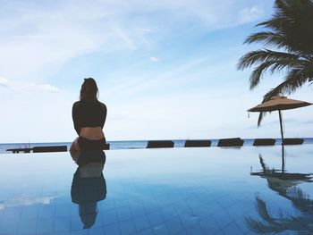 Rear view of thoughtful woman sitting by swimming pool against sky