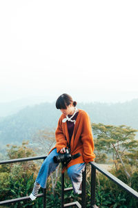 Young asian woman with casual clothes sit on fence in morning light holding camera at mountain top