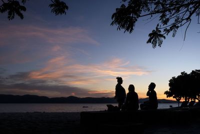 Silhouette people sitting on shore against sky during sunset