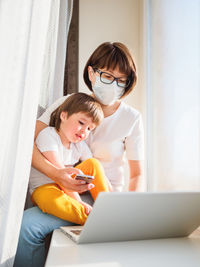 Woman in protective medical mask works remotely from home with laptop and toddler boy. coronavirus q