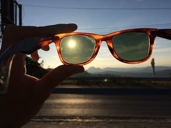 Close-up of hand holding sunglasses against sky during sunset