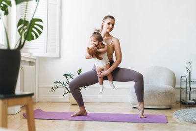 Young woman fit mom with baby girl in her arms doing fitness on mat at home