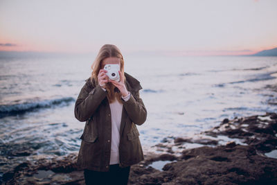 Portrait of young woman photographing sea against sky during sunset