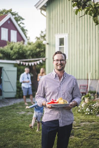 Portrait of smiling mature man holding food plate while standing in backyard
