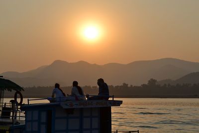 Silhouette people sitting on sea against mountains during sunset