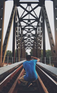 Rear view of man crouching by railroad tracks on bridge
